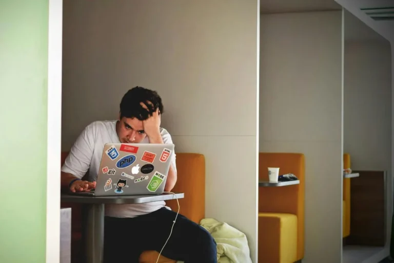 A stressed developer working on a laptop in a cafe booth symbolizes the challenges of entrepreneur addiction and the role of technology in overcoming it.