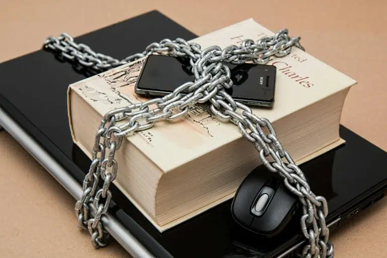 A laptop, book, phones, and mouse are bound by a heavy chain, symbolizing Blockchain and Cybersecurity as tools for building digital trust through secure access, data integrity, and protection against information restriction.