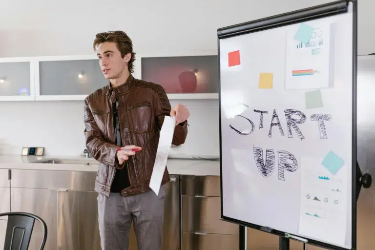 A presenter gestures beside a whiteboard labeled “START UP” with charts and sticky notes, symbolizing SEO for Fintech Startups as a strategy for building authority and driving growth through visibility and planning.