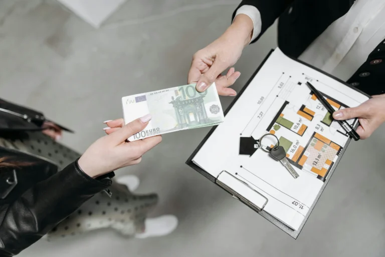 Two people exchange money, keys, and a floor plan during a property deal, symbolizing Real Estate in 2026 with market shifts, investment trends, and sustainability at the center.