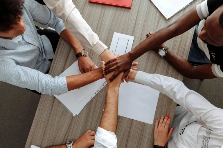Six employees join hands at a meeting table, symbolizing collaboration and unity—capturing the future of Employee engagement in 2026 through teamwork, shared purpose, and inclusive culture.
