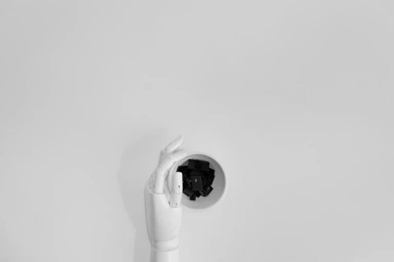 A white robotic hand holds a bowl filled with black keyboard keys against a plain background, symbolizing AI Transformation in the Industry through the fusion of robotics and digital interfaces, representing the shift toward automation, intelligent systems, and human-machine collaboration in modern industrial workflows.