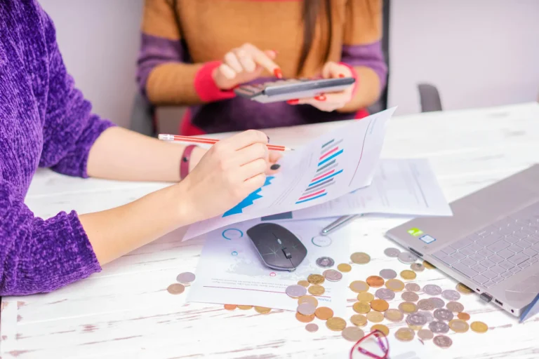 Two individuals analyze printed charts and use a calculator at a workspace scattered with coins, a laptop, and financial tools, symbolizing Algorithmic Stock Market Tools: Automating Growth and Precision through data-driven decision-making, real-time analytics, and collaborative financial modeling that supports smarter investing and scalable portfolio strategies.