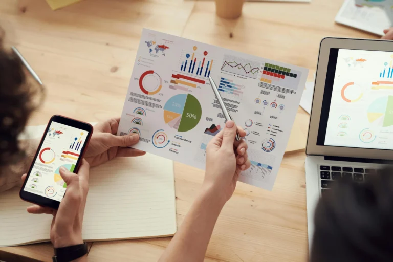 Professionals collaborate around synchronized data visualizations displayed on printed sheets, a smartphone, and a laptop, symbolizing Digital Transformation in Loans Automating Speed and Security through real-time analytics, cross-device integration, and performance metrics that drive faster loan processing, enhanced customer service, and secure financial operations.