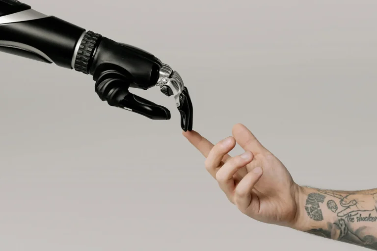 A robotic hand and a tattooed human hand reach toward each other with index fingers nearly touching, ymbolizing Emerging Tech in the Industry Driving Change through human-machine collaboration, advanced robotics, and the evolving interface between innovation and identity in modern industrial transformation.