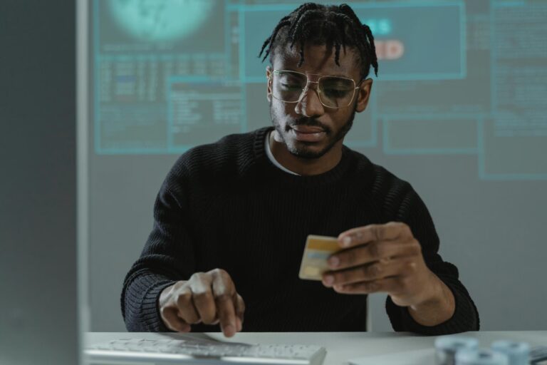 A person in glasses and a black sweater holds a credit card while interacting with a keyboard, with a digital interface of charts, data panels, and a world map in the background, symbolizing Fintech Funding Trends Shaping Digital Finance through secure online transactions, global data ecosystems, and investor-backed innovation driving the evolution of financial technology.