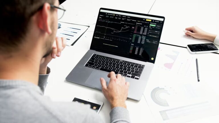 A person works at a desk analyzing cryptocurrency charts on a laptop, surrounded by printed graphs, a smartphone, and a tablet, symbolizing Fintech Investment Opportunities Driving Global Growth through data-driven decision-making, decentralized finance platforms, and digital tools that empower investors to capitalize on emerging global markets and scalable financial technologies.