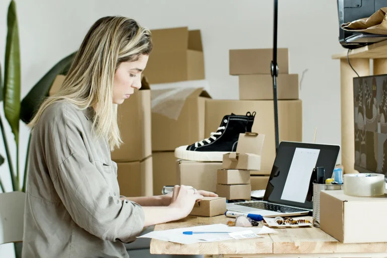 A person prepares packages at a workspace with cardboard boxes, a laptop, a sneaker, and shipping tools, symbolizing Fintech for Business Growth Driving Global Expansion through e-commerce logistics, digital payment integration, and scalable operations that empower entrepreneurs to streamline fulfillment and reach global markets.