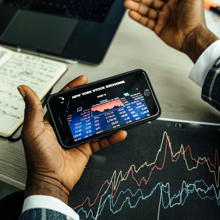 A suited individual holds a smartphone displaying real-time stock market indices from the New York Stock Exchange, with a laptop, handwritten notes, and printed financial charts in the background; symbolizing Future of Stock Market Technology Unlocking Advantage through mobile analytics, data visualization, and fintech tools that empower strategic decision-making and competitive edge in modern investing.