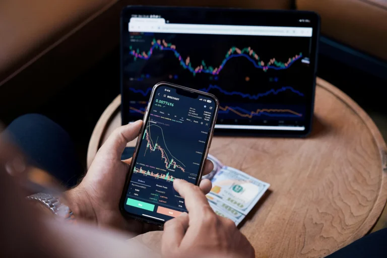 A hand holds a smartphone displaying a BIDZ/USDT crypto trading chart, with a tablet showing financial indicators and two $100 bills on a wooden desk, symbolizing Invest Money with Confidence Smart Wealth and Risk Strategies through real-time market analysis, diversified asset tracking, and digital tools that empower informed investment decisions and risk-managed financial growth.