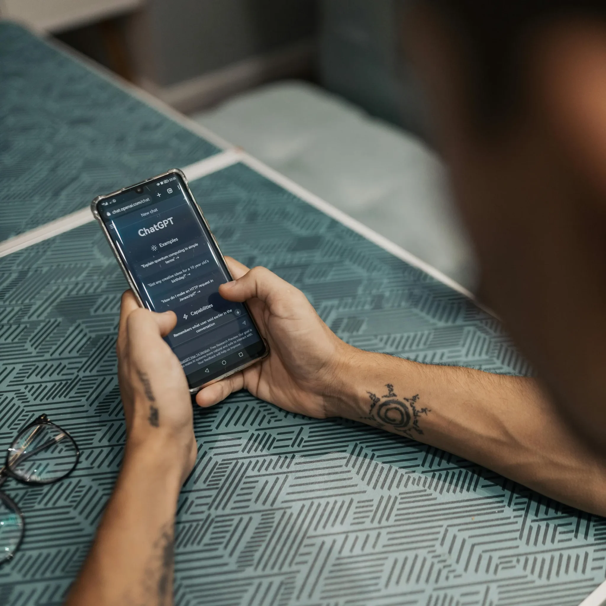 A person with tattooed forearms interacts with the ChatGPT interface on a smartphone at a patterned table, symbolizing OpenAI Outsourcing Automation for Business Growth through accessible AI tools, mobile integration, and streamlined support for entrepreneurs seeking scalable, intelligent solutions.
