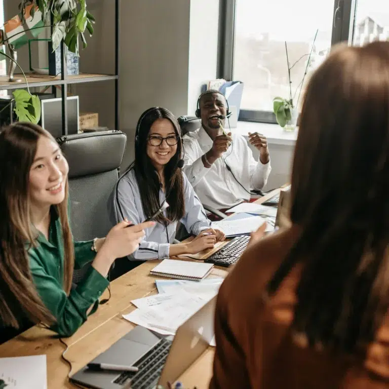 A diverse team collaborates around a table with laptops and headsets in a bright office, symbolizing SBA Support for Wellness Startups: Driving Growth through inclusive teamwork, digital tools, and strategic planning in health-focused ventures.