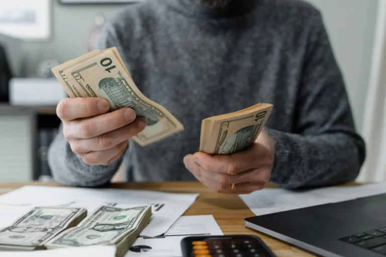 A person counts stacks of $10 bills at a desk surrounded by financial documents, a calculator, and a laptop, symbolizing Saving Plan Loan Automation for Smarter Growth through hands-on budgeting, digital finance tools, and strategic cash flow management that supports scalable lending solutions and optimized savings plans.