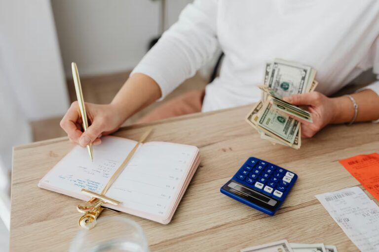 A person manages finances at a desk, holding U.S. dollar bills while writing in a planner labeled with expenses like rent, groceries, and savings. Surrounding items include a blue calculator, receipts, a smartphone, and a glass of water; symbolizing Saving Plan Loan Options Balancing Growth and Liquidity through hands-on budgeting, cash flow tracking, and strategic financial planning that supports both short-term needs and long-term asset growth.