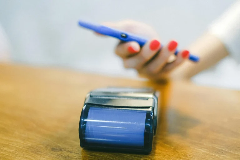 A person with red-painted nails holds a blue smartphone near a POS terminal on a wooden counter, initiating a contactless payment, symbolizing Startup Fintech Funding Driving Innovation and Growth through mobile-first financial solutions, seamless transaction experiences, and investor-backed technologies that empower agile startups to reshape the future of digital finance.