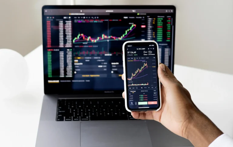 A person holds a smartphone displaying a BTC/USDT candlestick chart while monitoring a laptop filled with trading data; symbolizing Stock Market Automation Platforms Transforming Trading through real-time analytics, algorithmic execution, and synchronized interfaces that empower investors to make faster, data-driven decisions in dynamic crypto and equity markets.