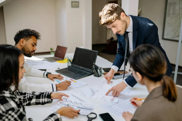 A group of professionals collaborate around a white table covered with charts, graphs, and laptops in a modern office; symbolizing Stock Market Discipline Driving Long Term Success through data-driven teamwork, strategic analysis, and consistent financial planning that supports sustainable portfolio growth and informed decision-making across evolving market conditions.