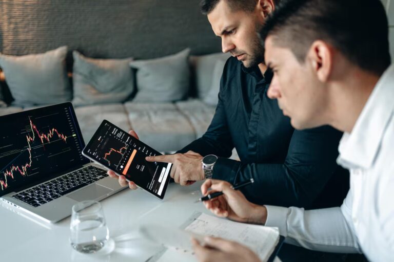 Two individuals collaborate in a modern setting using a tablet and laptop displaying stock market charts with candlestick patterns and trading data, while one takes notes, symbolizing Stock Market Fintech Transforming Trading and Analytics through real-time data visualization, mobile-first platforms, and collaborative decision-making that empower investors with smarter, faster market insights.