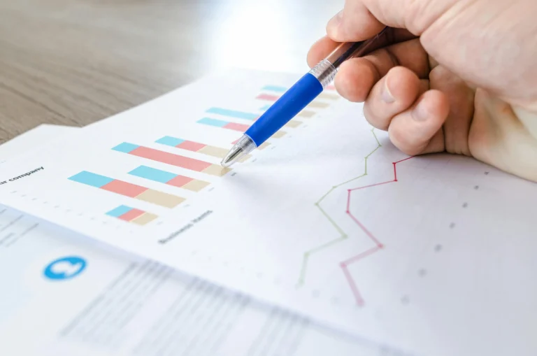 A close-up of a hand pointing with a pen at a printed document featuring colorful bar and line graphs; symbolizing Stock Market Growth Trends Patterns and Opportunities through hands-on data interpretation, visual trend analysis, and strategic insights that guide investment decisions and reveal market dynamics over time.