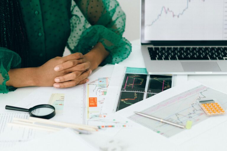 A person sits at a desk with hands clasped, surrounded by financial charts, a magnifying glass, calculator, and a laptop displaying a line chart; symbolizing Stock Market Research Driving Smarter Investment Decisions through technical analysis, data interpretation, and strategic tools that guide informed portfolio choices and market timing.