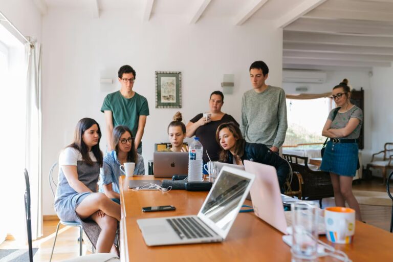 Eight individuals gather around a table in a modern, well-lit room, focused on a laptop screen amid coffee mugs, notebooks, and tech devices, symbolizing Stock Market Trends Shaping Investor Strategies through collaborative analysis, real-time data engagement, and group-driven decision-making that reflect how investors adapt to evolving market signals and strategic insights.