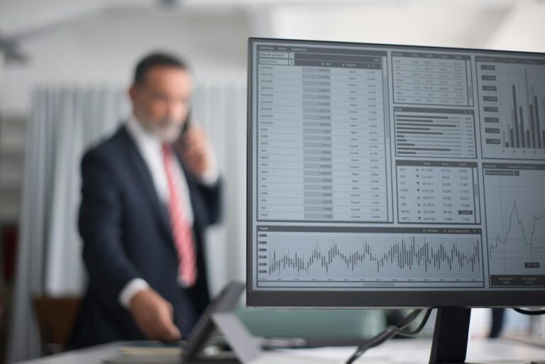A financial trading dashboard on a computer monitor displays charts, tables, and profit metrics, while a blurred professional in a business suit speaks on the phone, symbolizing Stock Trading Investments for Wealth Building through real-time market analysis, strategic portfolio management, and technology-driven decision-making in professional financial environments.