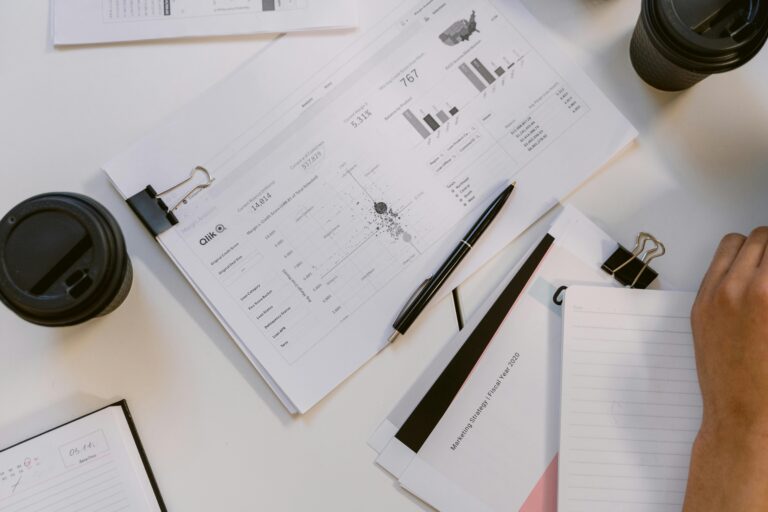 A workspace features Qlik data reports with charts, bar graphs, and a U.S. map showing regional metrics like “Conversion Rate” and “Customer Count,” alongside coffee cups and a hand reviewing documents, symbolizing US Bank Marketing Analytics through performance dashboards, fiscal strategy insights, and data-driven decision-making that optimize customer acquisition and regional growth initiatives.