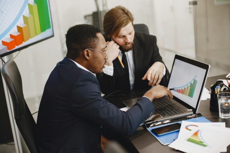 Two professionals in business attire analyze a laptop screen displaying a colorful upward-trending bar graph, with similar growth visuals on a monitor and printed documents, symbolizing AI Business Growth in the Digital Economy through data-driven strategy, collaborative decision-making, and intelligent forecasting that empower founders and teams to scale operations, optimize performance, and lead innovation in tech-enabled markets.
