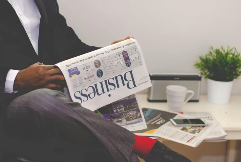 A person in business attire reads an upside-down newspaper titled “Business” while seated in an office space with a desk holding a potted plant, coffee mug, smartphone, and other papers, symbolizing Economic Effects on Industries: News and Insights through a blend of market headlines, global index references, and casual professional observation that reflect how founders and analysts interpret economic signals, sector shifts, and financial media narratives in real time.