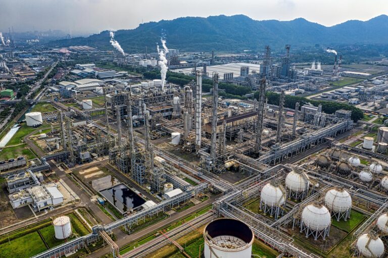 Aerial view of a sprawling industrial refinery complex with distillation towers, piping systems, and spherical tanks emitting steam, symbolizing Economic Forecast Reports for Global Growth through energy production, infrastructure scale, and industrial output that influence macroeconomic indicators, global supply chains, and regional development trends.