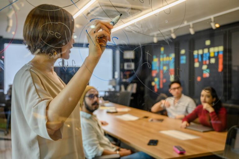 A person writes on a transparent glass board covered in circular diagrams while three others observe from a long table, with colorful sticky notes arranged on the wall behind, symbolizing Fintech Funding Strategies for Scalable Finance through collaborative planning, visual ideation, and agile workflows that support innovative financial models and scalable digital solutions.