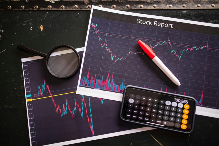 A financial analysis workspace features printed stock charts labeled “Stock Report,” a magnifying glass, a red and white pen, and a smartphone calculator displaying “100,365”, symbolizing From Hustle to Harmony: The 2026 Stock Trading Mindset through focused analysis, mindful investing, and strategic clarity that shift traders from reactive hustle to data-driven harmony in navigating modern financial markets.