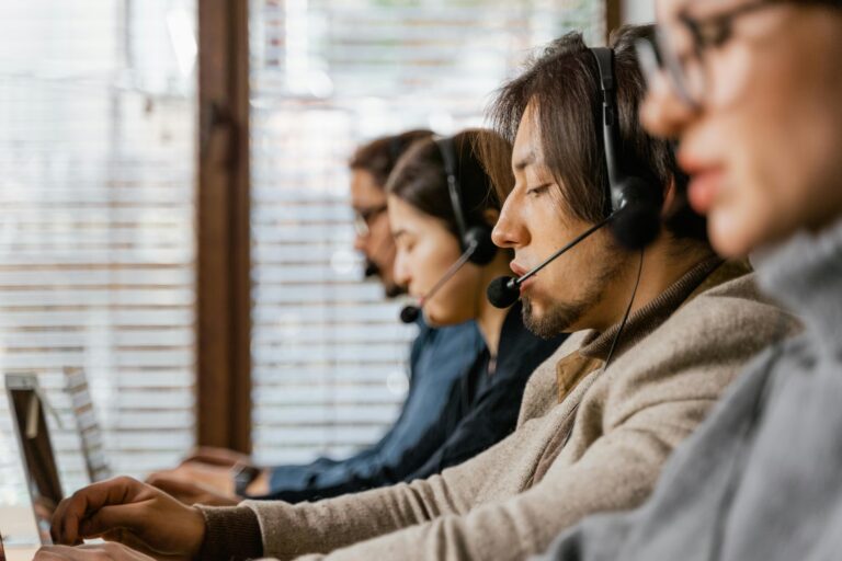 A row of professionals wearing headsets work on laptops in a brightly lit office with window blinds, suggesting a collaborative offshore support environment, symbolizing Global Industry Offshore Outsourcing Insights through remote workforce efficiency, cross-border service delivery, and scalable enterprise operations that empower founders and global firms to optimize costs, enhance customer experience, and expand capabilities through strategic outsourcing.