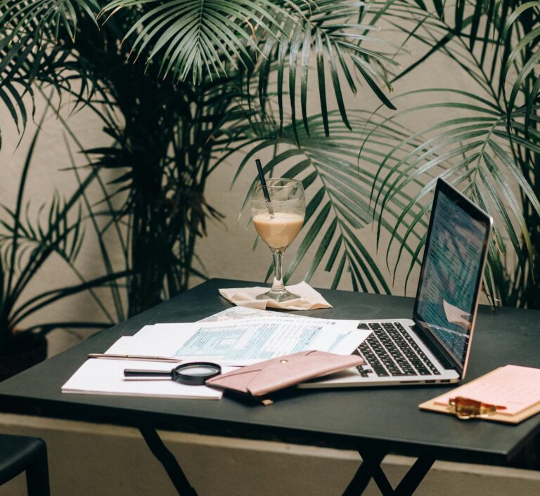 A workspace setup on a black table features a laptop displaying a spreadsheet, printed documents, a magnifying glass, a pink wallet, a clipboard with a pink notepad, and a glass of iced coffee, symbolizing Industry Search Visibility Tips for the 2026 Agentic Era through data-driven content analysis, mobile-friendly optimization, and strategic keyword mapping in a serene, nature-infused environment that reflects the evolving blend of productivity, personalization, and ambient intelligence.