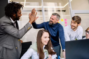 Five professionals celebrate in a modern office, with two giving a high-five while others smile and engage near a laptop and a wall of colorful sticky notes, symbolizing Jobs Report Workplace Satisfaction and Growth through team camaraderie, collaborative success, and founder-led culture strategies that empower organizations to boost employee morale, align productivity with purpose, and drive sustainable business expansion in dynamic work environments.
