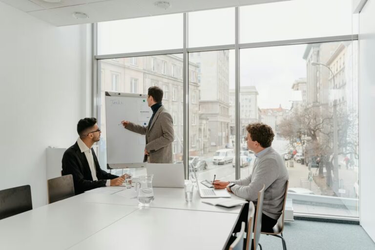 A professional leads a strategy session in a modern office, writing “Tasks:” on a flip chart while two colleagues observe and take notes, symbolizing Leadership in the Era of Industry Global Workforce Trends through collaborative planning, adaptive management, and cross-functional engagement that reflects evolving workplace dynamics, global talent integration, and strategic leadership in a connected economy.
