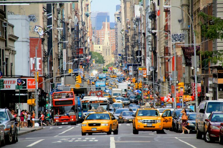 A bustling New York City street filled with yellow taxis, buses, and pedestrians, framed by dense urban architecture and a rising church spire, symbolizing Mastering Economic Conditions for Systemic Growth in 2026 through metropolitan activity, infrastructure intensity, and the dynamic interplay of commerce, mobility, and cultural resilience that shape macroeconomic expansion and urban-driven innovation.