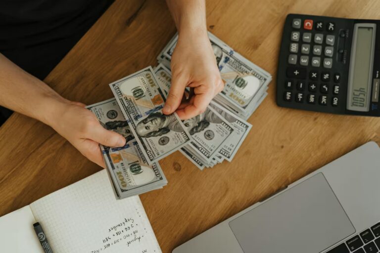 A person counts a stack of U.S. $100 bills on a wooden table surrounded by a calculator displaying “1054,” a laptop, and a handwritten budget notebook, symbolizing Mastering Growth with Saving Plan Loan Tools through disciplined financial tracking, strategic budgeting, and loan-supported savings plans that empower individuals and businesses to build capital and scale responsibly.