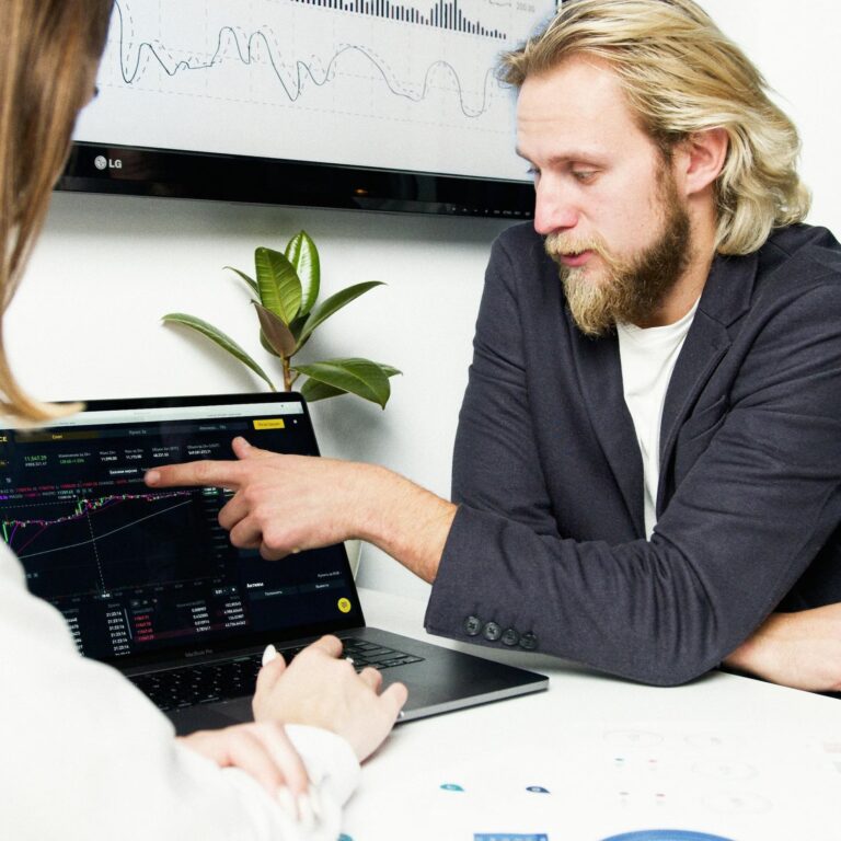 Two individuals analyze financial charts on a laptop and large monitor in a modern workspace, symbolizing Navigating the 2026 Global Economic Trends through collaborative market analysis, data-driven forecasting, and strategic insights into global financial performance, investment behavior, and macroeconomic shifts.