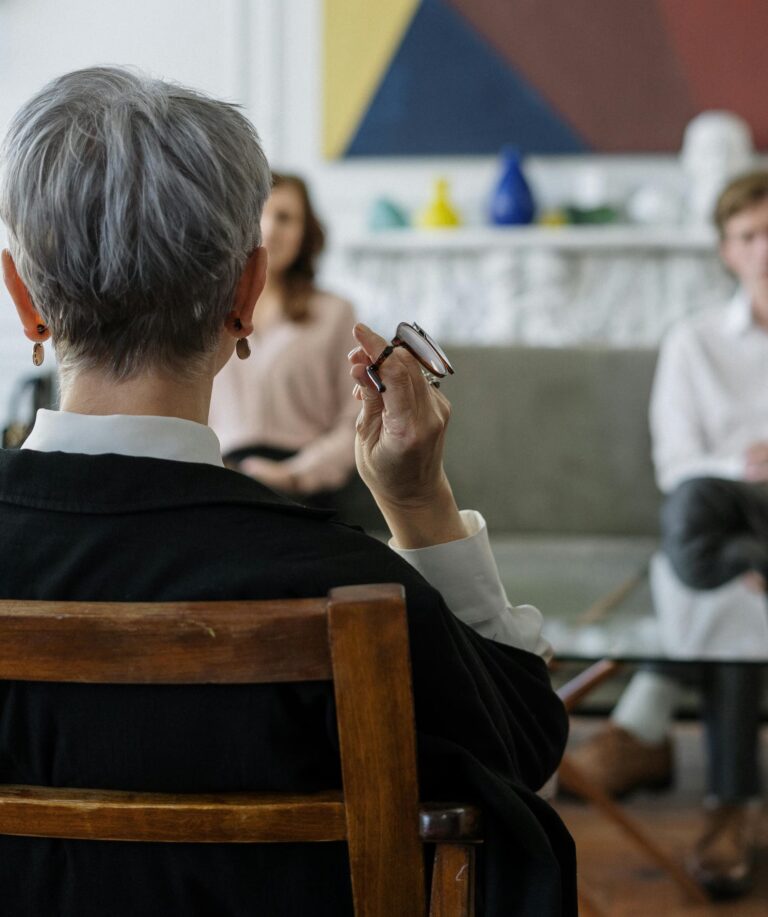 An older professional sits across from two individuals in a modern, well-decorated room, holding eyeglasses and engaging in a focused conversation, symbolizing Practical Stock Trading Tips for Confident Investing through mentorship, strategic dialogue, and personalized guidance that empowers investors to navigate market trends, manage risk, and build confidence in their trading decisions.