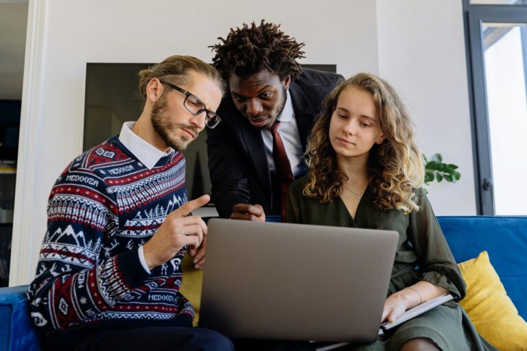 Three individuals collaborate around a laptop in a modern workspace, with one pointing at the screen, another holding a notebook, and a third leaning in attentively, symbolizing SBA Loan Programs for Startup Success through teamwork, digital planning, and strategic guidance that support entrepreneurs in launching, funding, and growing new ventures.