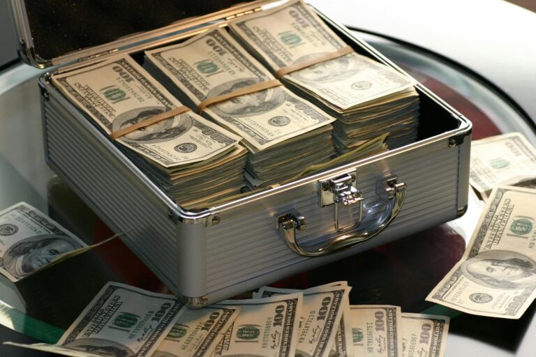 An open metallic briefcase filled with neatly bundled stacks of U.S. $100 bills sits on a glass table, with additional loose bills scattered around, symbolizing Scale Success with U.S Bank Business Funding through accessible capital, structured financing, and strategic liquidity that empower businesses to expand operations, invest confidently, and accelerate growth in competitive markets.
