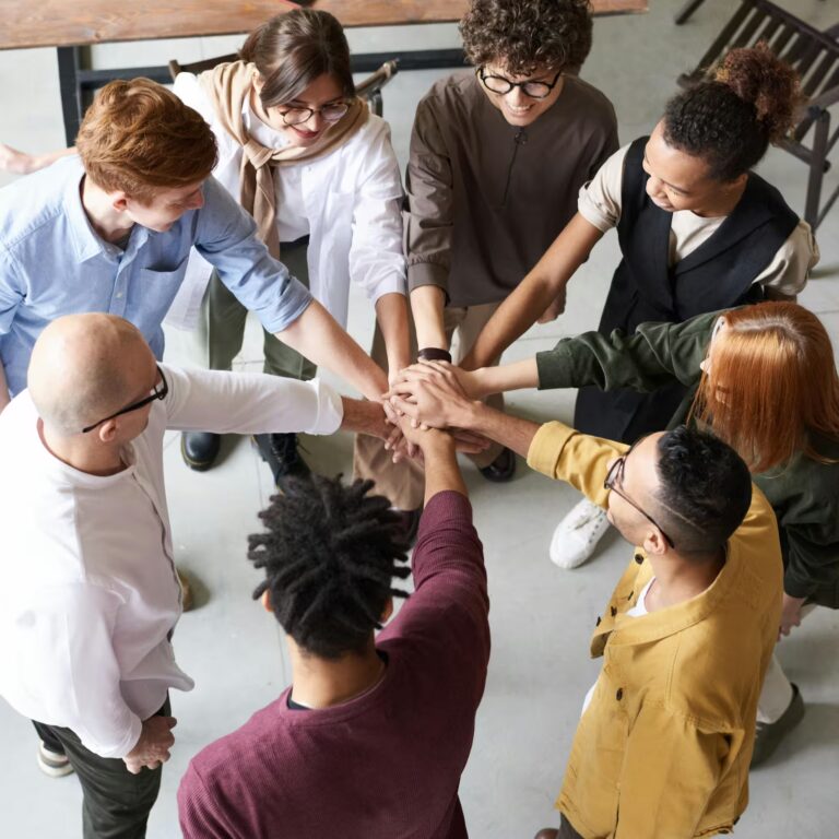 Eight individuals stand in a circle with hands stacked at the center in a gesture of unity, symbolizing Scale Using Global Talent in the Industry through collaborative teamwork, inclusive culture, and cross-border synergy that drives innovation, operational growth, and industry-wide scalability.