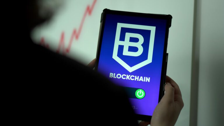 A person holds a tablet displaying a blue screen with a shield logo containing the letter “B” and the word “BLOCKCHAIN” beneath it, with a green power icon at the bottom and a blurred red financial graph in the background, symbolizing Scaling On-Chain with Blockchain Fintech Solutions through secure digital infrastructure, decentralized transaction systems, and scalable fintech innovation that empower founders to build resilient, trustless financial ecosystems.
