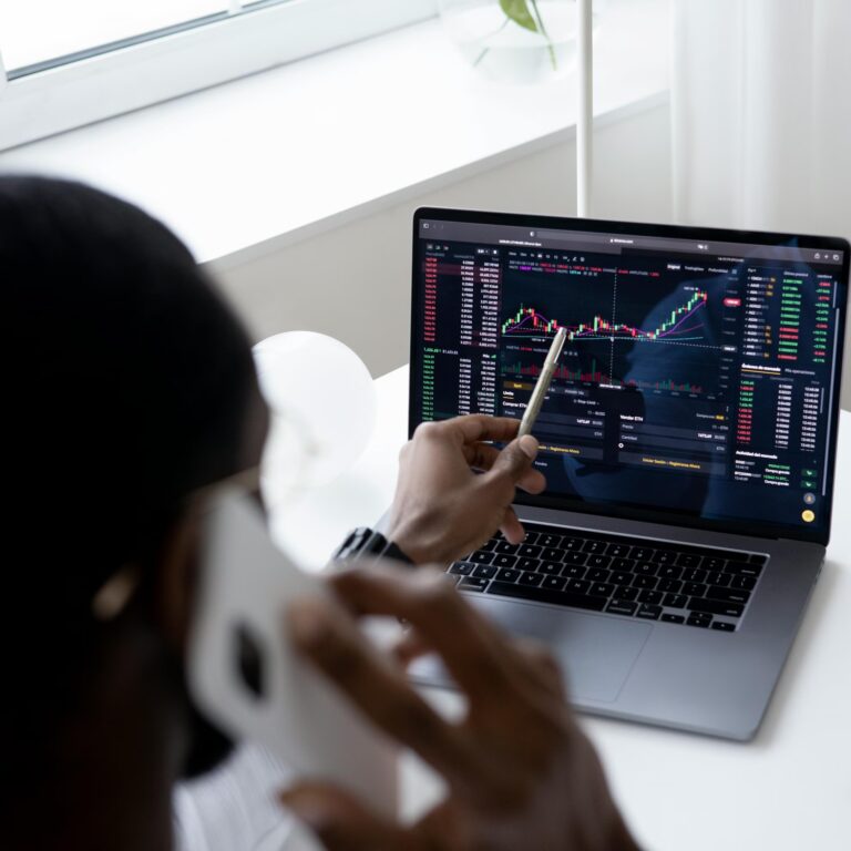 A person actively engages in financial trading, holding a smartphone while pointing at a laptop screen filled with candlestick charts, market data, and price movements, symbolizing Scaling Success in the Global Stock Market through real-time analysis, tech-enabled decision-making, and strategic positioning across international equities and digital assets.