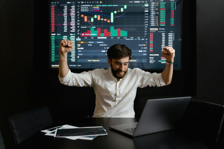 A person raises both arms in celebration at a desk with a laptop, while a large screen behind displays a cryptocurrency trading interface filled with candlestick charts, market data, and trading pairs, symbolizing Stock Trading Discipline for Long-Term Success through strategic execution, emotional control, and founder-led analysis that empower investors to navigate volatility, apply consistent methods, and build resilient portfolios in dynamic financial markets.
