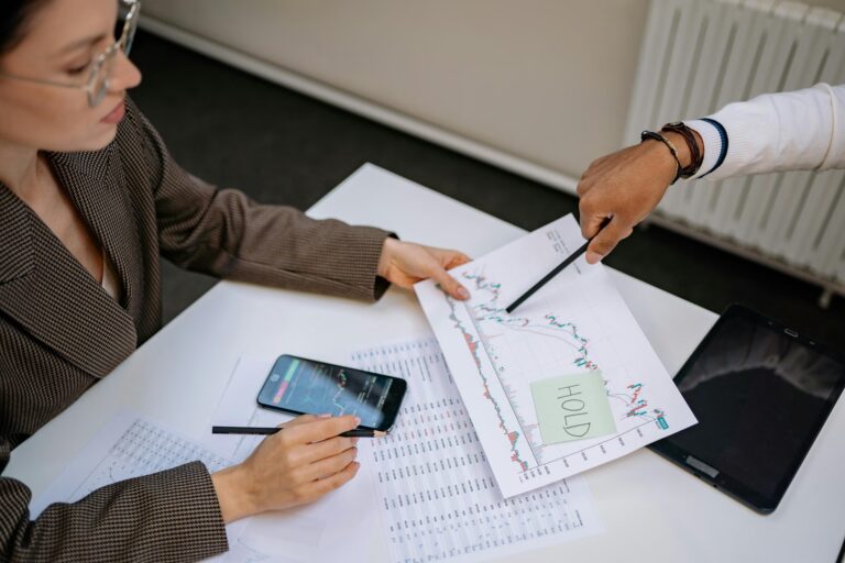 Two individuals review a printed stock chart with candlestick patterns and a sticky note labeled “HOLD,” while referencing data on a smartphone and spreadsheets, symbolizing Stock Trading Guide for Confident Market Participation through collaborative analysis, strategic decision-making, and multi-device trading insights that empower investors to act with clarity, discipline, and market confidence.