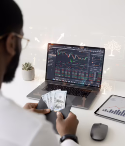 A person holds several U.S. hundred-dollar bills in an envelope while analyzing financial trading charts on a laptop, with a tablet displaying bar and line graphs nearby, symbolizing Stock Trading for Expansion and Wealth Building through active market analysis, liquidity management, and founder-led investment strategies that empower individuals to scale capital, seize growth opportunities, and build long-term wealth in dynamic trading environments.