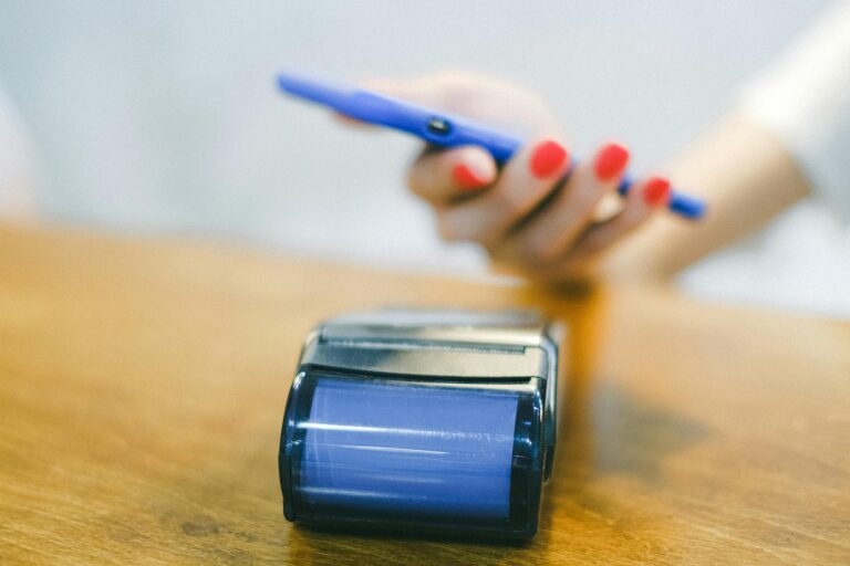 A person holds a blue smartphone near a payment terminal on a wooden surface, initiating a contactless transaction, symbolizing The Ecosystem Engine Integrated Fintech Platforms through seamless digital payments, mobile-first financial tools, and interconnected systems that drive real-time commerce, user-centric innovation, and scalable fintech infrastructure.