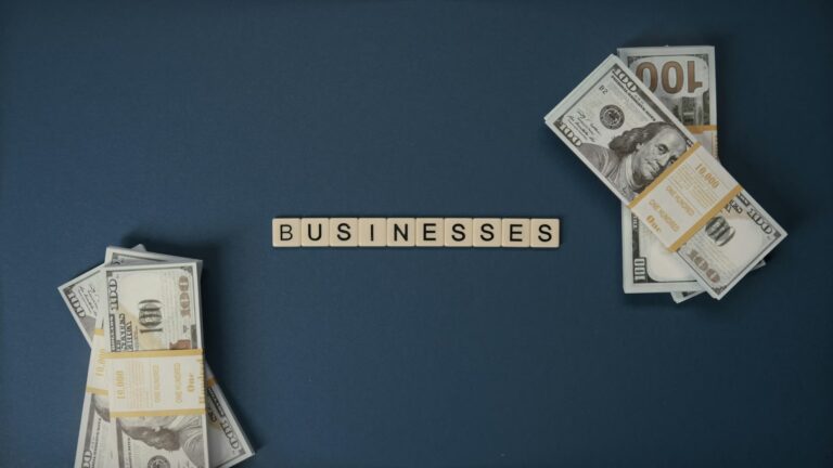 Stacks of U.S. $100 bills arranged on a dark blue surface with beige letter tiles spelling “BUSINESSES”, symbolizing Working Capital Loans as Efficiency Multipliers through liquidity access, operational funding, and strategic capital deployment that accelerates business productivity, cash flow stability, and scalable growth.